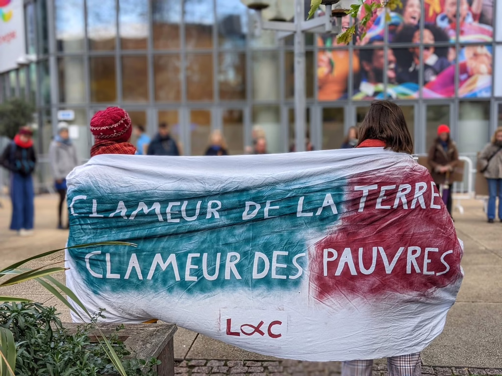 deux militant.e.s de Lutte et Contemplation de dos portent une banderole marquée "Clameur de la Terre et Clameur des Pauvres, Luc" en lettres blanches sur des calques bleus et rouges