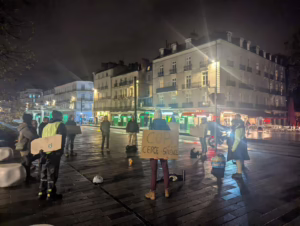 Des militant.e.s de Lutte et contemplation sont debout, en cercle, en silence, sur une place animée, en plein hiver. Au premier plan, un des militants tient une pancarte "COP29, cercle, silence"